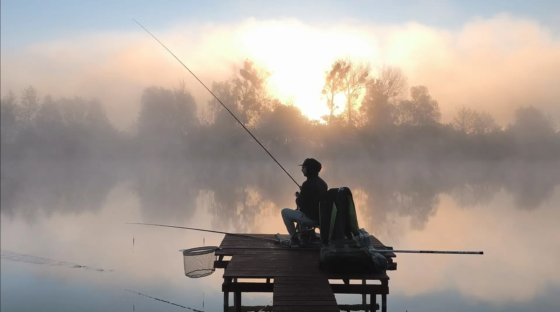 Wędkarz siedzący na drewnianym pomoście podczas mglistego wschodu słońca nad spokojnym jeziorem